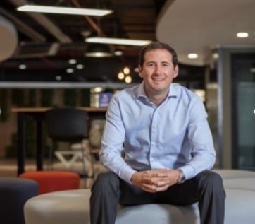Smiling man in a light blue shirt sits on a round gray ottoman in a modern office lounge area. - Las poderosas familias peruanas que hacen fortuna en Colombia con gaseosas, colegios y cosméticos