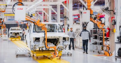 Automotive assembly line with a white car body on a yellow platform and orange robotic arms overhead in a bright factory. - Las marcas de carros que mueven sus hilos en Suramérica y buscan montar sus plantas cerca a Colombia