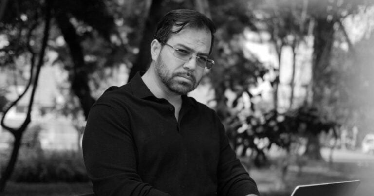Bearded man wearing glasses and a black polo, working on a laptop outdoors in a park setting. - Dios no hace campaña