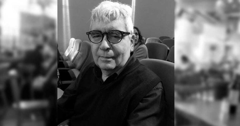 Older man with short white hair and glasses seated in a row of theater chairs, facing the camera. - La trampa de Tucídides