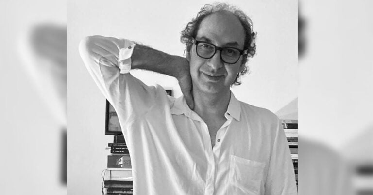 Man with glasses in a white button-down shirt, posing with his arm behind his neck indoors. - La odisea de aguantar la odisea