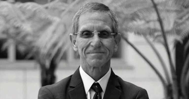 Older man in a suit and tie wearing glasses, outdoors with blurred palm plants in the background (black and white).] , - Marcas, patentes, competencia y consumidores