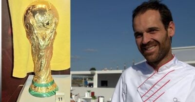 Gold FIFA World Cup trophy on a pedestal with a green base, next to a smiling male chef in a white uniform outdoors. - Tras la conquista del Mundial, así celebra la comunidad española en Bogotá