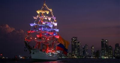 Tall sailing ship illuminated with multicolored lights at night, flying a Colombian flag, with a city skyline in the background. - Las2orillas.co: Historias, voces y noticias de Colombia