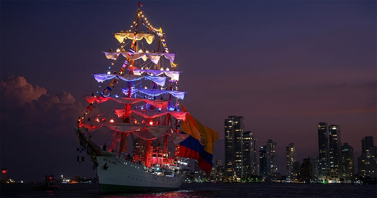 Tall sailing ship illuminated with multicolored lights at night, flying a Colombian flag, with a city skyline in the background. - El ARC Gloria y su entrada en Nueva York como embajador del país en los 250 años de la independencia de Estados Unidos