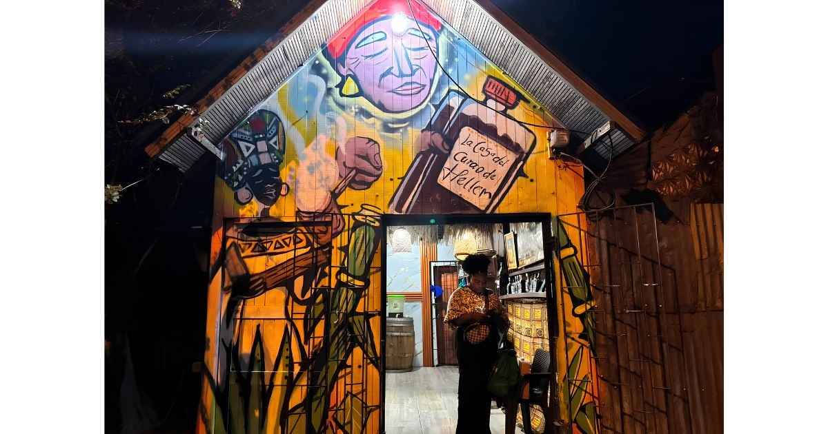 Una mujer en Tumaco logró llevar su viche ancestral a grandes escenarios como el Petronio Álvarez, la COP 16 y Anato Nighttime view of a graffiti-covered building with a doorway; a person stands inside near shelves and barrels, framed by bright mural art with fists and faces. - Una mujer en Tumaco logró llevar su viche ancestral a grandes escenarios como el Petronio Álvarez, la COP 16 y Anato