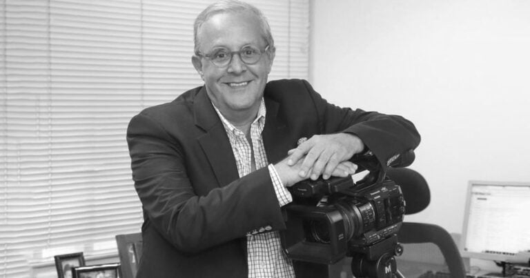 Smiling man in a blazer with glasses rests his hands on a professional video camera on a tripod in an office setting. - Por el bien de Colombia, el Valle debe ser una prioridad para Abelardo