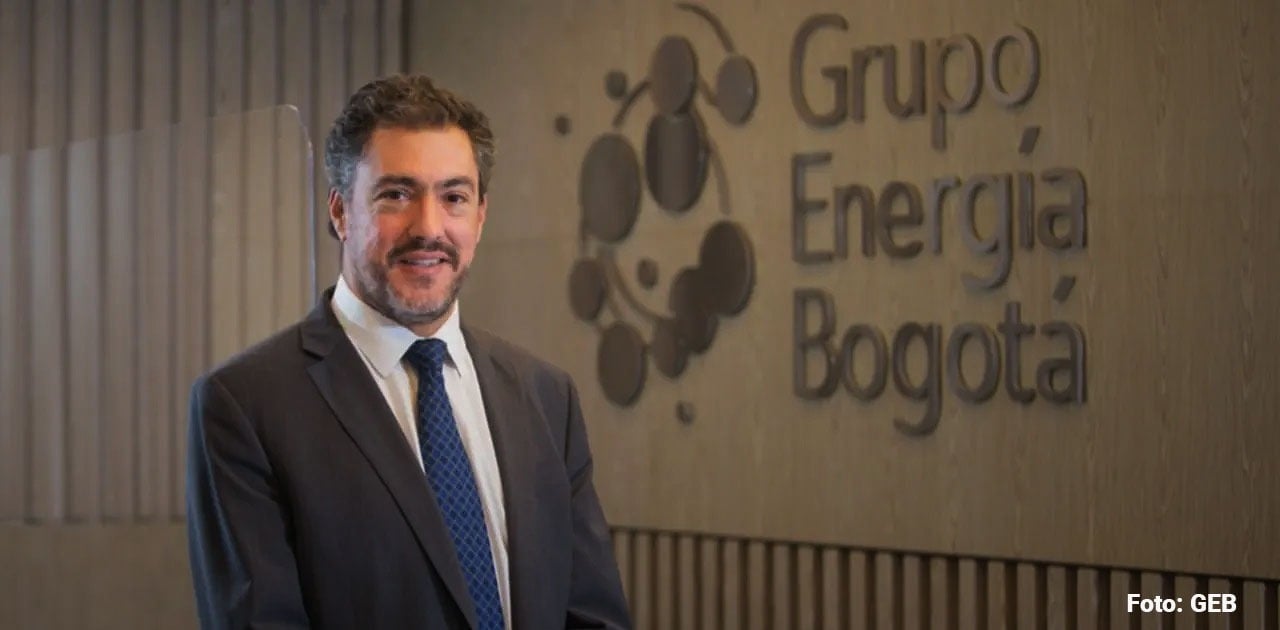 Man in a dark suit and blue tie stands in a hallway beside a textured wall with the Grupo Energía Bogotá logo in the background. - Juan Ricardo Ortega y su poderosa junta convirtieron al Grupo Energía de Bogotá en un poderoso jugador en elBrasil