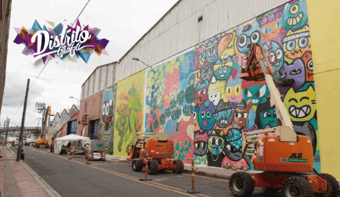 Un recorrido por las historias que el aerosol escribió en los muros de Bogotá: Grafiti Tour Colorful graffiti mural along a city street on a large building, with orange cherry-pickers and workers; Bogotá D.C. District Graffiti logo in the upper left. - Un recorrido por las historias que el aerosol escribió en los muros de Bogotá: Grafiti Tour