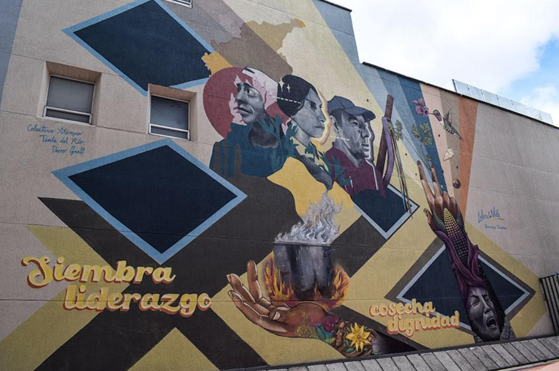 Un recorrido por las historias que el aerosol escribió en los muros de Bogotá: Grafiti Tour Colorful mural on a building wall with geometric shapes, multiple portraits, and a hand holding a flame among bold colors. - Un recorrido por las historias que el aerosol escribió en los muros de Bogotá: Grafiti Tour