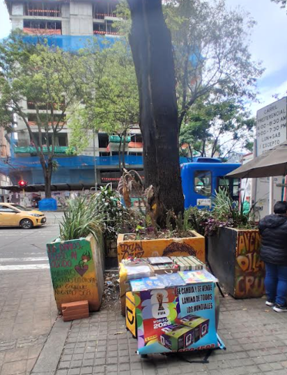 Un recorrido por las historias que el aerosol escribió en los muros de Bogotá: Grafiti Tour Urban sidewalk scene with a large tree in colorful planter boxes, a vending/newsstand with posters, and a blue bus in the background. - Un recorrido por las historias que el aerosol escribió en los muros de Bogotá: Grafiti Tour