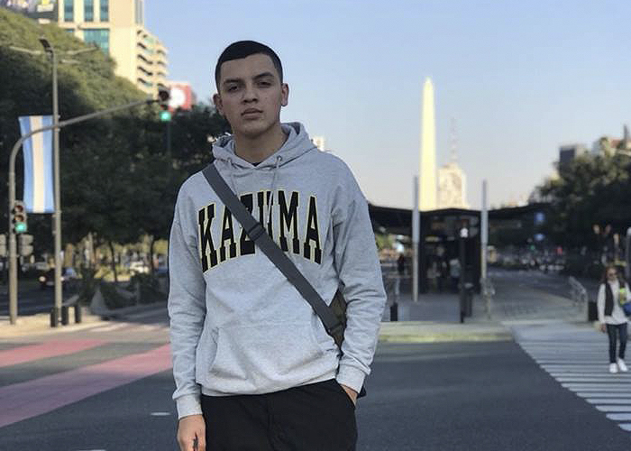 Un recorrido por las historias que el aerosol escribió en los muros de Bogotá: Grafiti Tour Young man wearing a gray hoodie with a crossbody bag, standing on a city street near a crosswalk. - Un recorrido por las historias que el aerosol escribió en los muros de Bogotá: Grafiti Tour