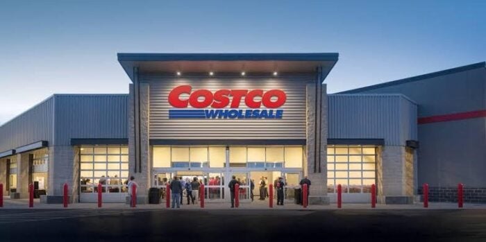 El negociazo con el que el empresario paisa Santiago Botero busca conquistar a EE. UU. Costco Wholesale storefront with illuminated red and blue sign, shoppers lined up at the entrance at dusk. - El negociazo con el que el empresario paisa Santiago Botero busca conquistar a EE. UU.