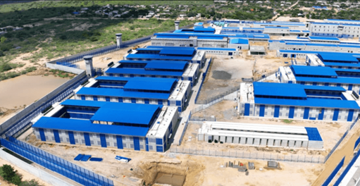 Sitio de construcción con muchos edificios rectangulares de techo azul dispuestos en filas, perímetro cercado.