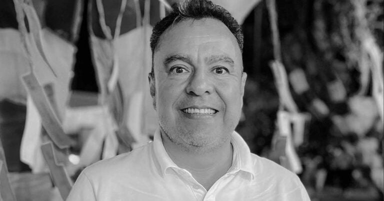 Smiling man in a white shirt facing the camera at a festive event with streamers in the background (black-and-white). - País y regiones sin espejo