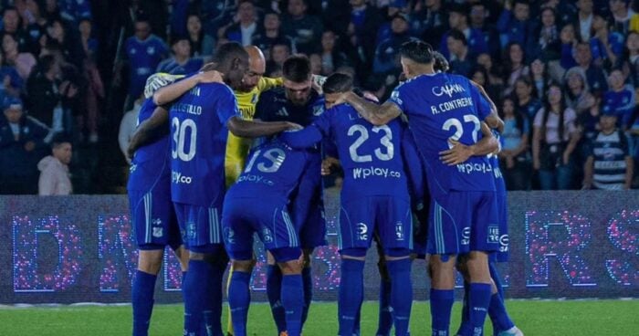 El francés dueño Caracol Radio quiere sacarle una fortuna a Millonarios, el equipo bogotano que compró hace 15 años Blue-clad soccer players form a huddle on the field, arms around each other, with fans in the stands behind them. - El francés dueño Caracol Radio quiere sacarle una fortuna a Millonarios, el equipo bogotano que compró hace 15 años