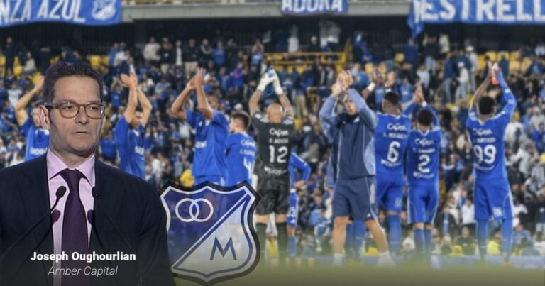 Las2orillas.co: Historias, voces y noticias de Colombia Man in a suit and glasses at a podium speaking in a stadium, blue-clad players celebrate behind him. - Las2orillas.co: Historias, voces y noticias de Colombia