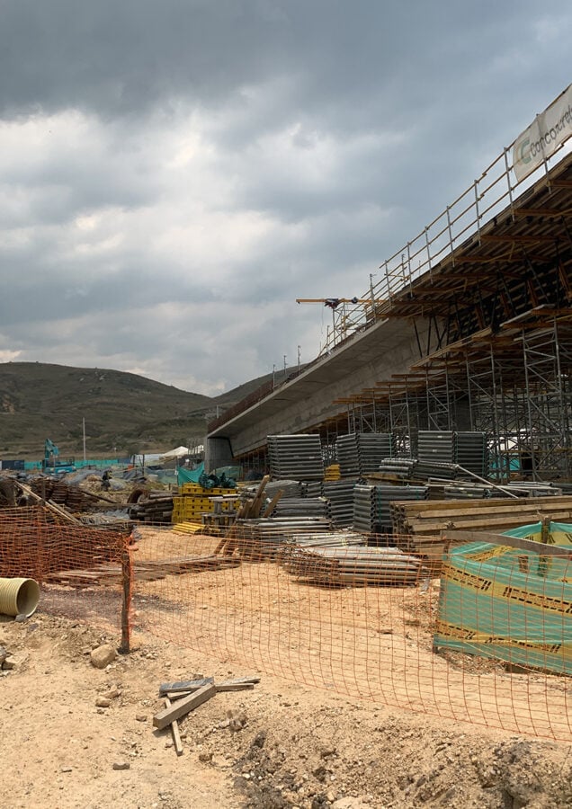 Bridge under construction on scaffolding with metal pipes and materials scattered on a dirt site under cloudy skies, hillside in the background.”], - Los Lozada aliados a una empresa francesa se quedaron con la billonaria licitación para los buses de Transmilenio