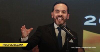 Male speaker in a dark suit and blue tie delivers a talk at a microphone; yellow - Las2orillas.co: Historias, voces y noticias de Colombia