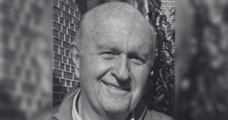 Smiling older man with short light hair in a grayscale photo, standing beside a brick wall. - No basta con cambiar de gobierno
