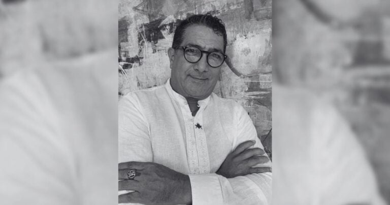 Portrait of a middle-aged man with glasses and a white kurta, arms crossed, posed against an abstract textured background (black and white photo). - Opinión impopular en tragedia