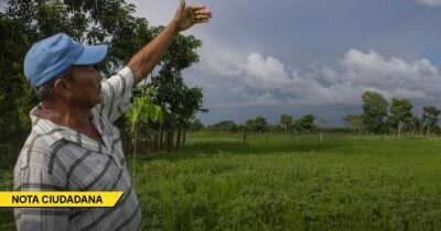 Man in a light plaid shirt and blue cap raises his right arm in a green rural field with trees and a cloudy sky; a yellow banner at the bottom left reads NOTA CIUDADANA. - Las2orillas.co: Historias, voces y noticias de Colombia