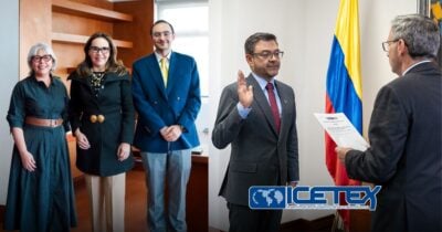 Four professionals stand together in an office; on the right, a man raises his right hand as another official reads from a document beside a Colombian flag, with an ICETEX logo in the corner. - Las2orillas.co: Historias, voces y noticias de Colombia