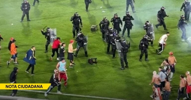 Riot police in black armor with shields confront a crowd of civilians on a grassy field, with a yellow Nota Ciudadana banner in the corner and smoke in the air. - El negociazo de transformar hinchas en vándalos dispuestos a morir por una camiseta