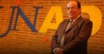 Smiling man in a suit posing in front of a brick wall with oversized blue and yellow letters behind him. - Tras el empujón de Petro a la UNAD, Jaime Leal se atornilló en la Rectoría otros 4 años: ya completa 27