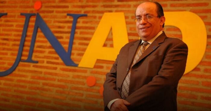 Smiling man in a suit posing in front of a brick wall with oversized blue and yellow letters behind him. - Las2orillas.co: Historias, voces y noticias de Colombia