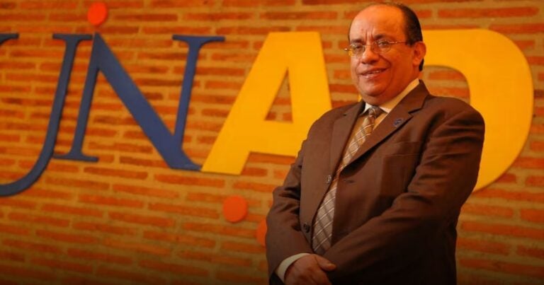 Smiling man in a suit posing in front of a brick wall with oversized blue and yellow letters behind him. - Tras el empujón de Petro a la UNAD, Jaime Leal se atornilló en la Rectoría otros 4 años: ya completa 27