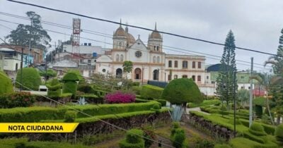 White church with twin domes behind a formal, sculpted garden and overhead utility lines under a cloudy sky; a yellow 'NOTA CIUDADANA' banner is in the lower-left corner. - Las2orillas.co: Historias, voces y noticias de Colombia