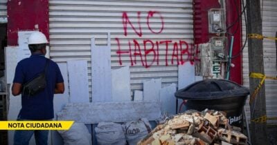 Construction worker in a white hard hat faces a shuttered storefront with red graffiti reading 'NO HABITABLE', debris and rubble in front. - Las2orillas.co: Historias, voces y noticias de Colombia
