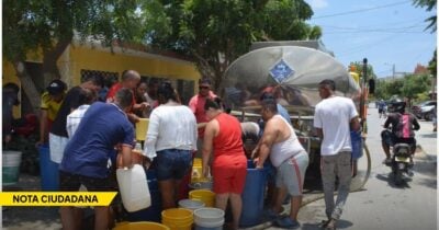 Community members gather around a water tanker on a sunny street, filling yellow and gray buckets as a crowd forms around the truck with a yellow banner reading 'NOTA CIUDADANA' visible. - Las2orillas.co: Historias, voces y noticias de Colombia