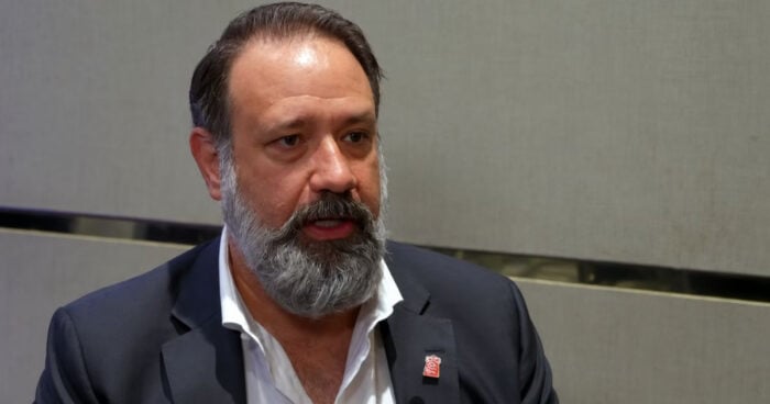 Quiénes son los hombres del lobby con los que el gobierno y los empresarios se mueven en Washington Middle-aged man with gray beard in a navy suit and white shirt, speaking in a conference room setting with a gray wall background, pin on his lapel visible. - Quiénes son los hombres del lobby con los que el gobierno y los empresarios se mueven en Washington