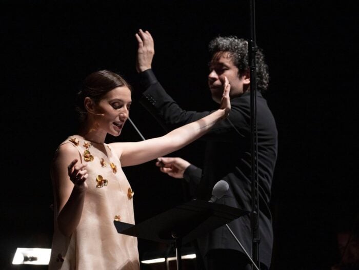 La envidiable historia de amor del director sinfónico venezolano Gustavo Dudamel y la actriz española María Valverde Woman in a pale dress with gold butterfly appliqués raises her arms on stage, headset mic engaged; a man in dark clothes claps in the background against black lighting.