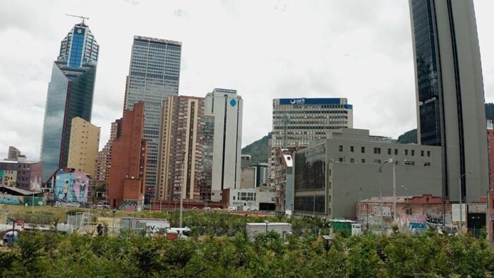 City skyline with tall modern buildings, a construction crane on a tower, graffiti walls, and a green foreground park area while traffic hums below. - Después de 13 años, el ambicioso Plan de Galán para el centro vuelve a ponerse en marcha