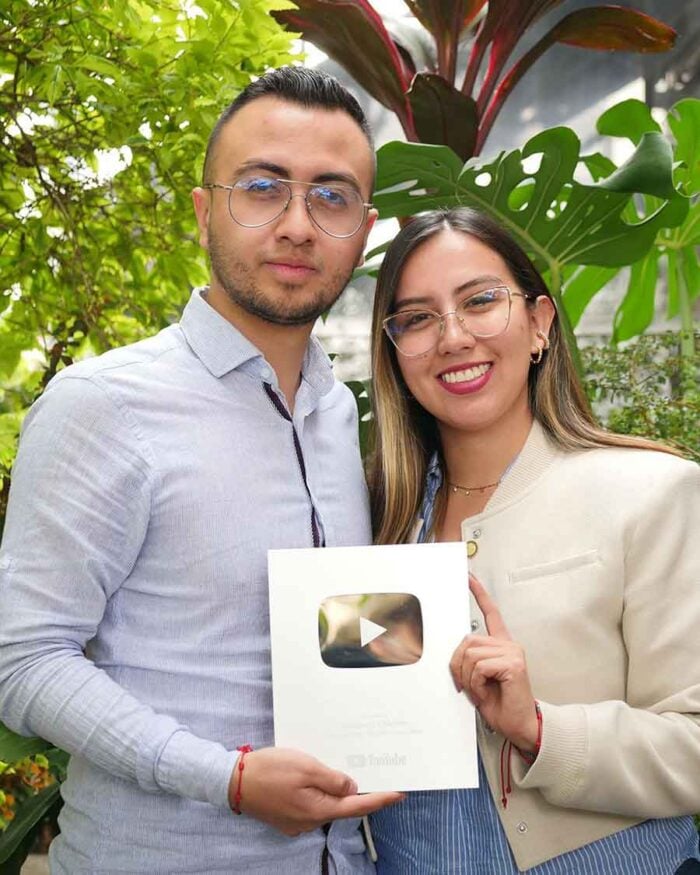 Así conquistaron dos periodistas colombianos el primer lugar de Spotify con historias de crimen Two people posing outdoors while holding a YouTube Creator Award plaque in front of green plants and big leaves. - Así conquistaron dos periodistas colombianos el primer lugar de Spotify con historias de crimen
