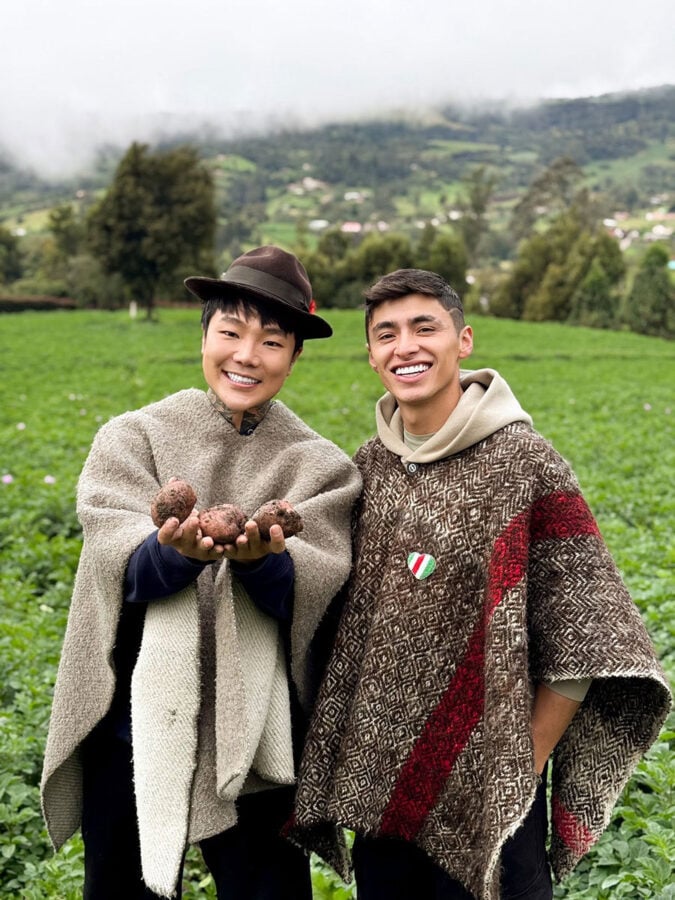 El influencer extranjero radicado en Colombia que es el número uno con 15 millones de seguidores en Instagram y TikTok Two smiling friends in a green field hold freshly harvested potatoes toward the camera, wearing thick ponchos with a rural hillside backdrop. - El influencer extranjero radicado en Colombia que es el número uno con 15 millones de seguidores en Instagram y TikTok