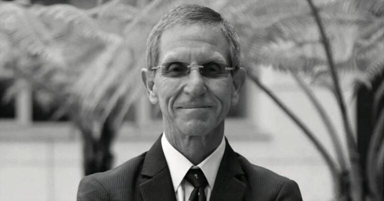 Smiling man in a dark suit and tie wearing glasses, outdoors with blurred plants in the background. - Registro Único de Proponentes para el Estado