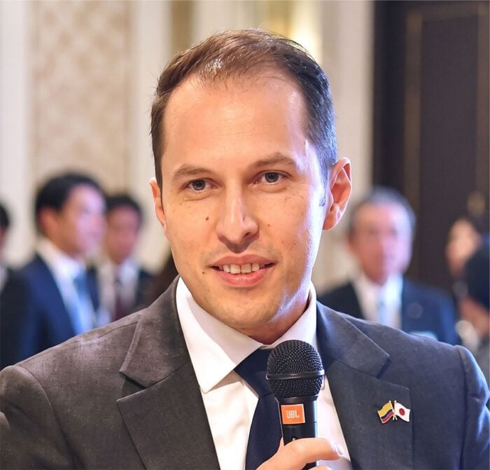 Quiénes están en los mejores oficinas de Procolombia en el exterior que El Tigre va a acabar Man in a dark suit speaks into a handheld microphone at a formal event, with blurred attendees in the background and two lapel pins on his jacket. - Quiénes están en los mejores oficinas de Procolombia en el exterior que El Tigre va a acabar