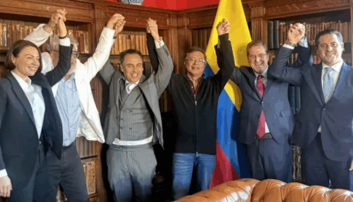 Quiénes son los hombres del lobby con los que el gobierno y los empresarios se mueven en Washington Group of six professionals in suits raise their clasped hands in celebration in a wood-paneled room with a Colombian flag in the background. - Quiénes son los hombres del lobby con los que el gobierno y los empresarios se mueven en Washington
