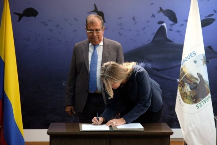 Tras 20 años dedicada a salvar una isla remota del Pacífico, la bióloga Sandra Bessudo cuidará los 65 Parques Nacionales A woman signs a document on a wooden desk as a suited man observes in a ceremony with flags and a marine backdrop behind them.