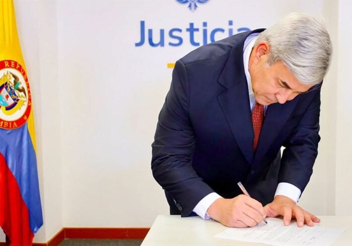 Por orden del presidente Dela Espriella, Joaquín Méndez superintendente de Notariado Bloqueó entrega de notarías Public official in a dark suit signs a document at a white table, Colombia flag to the left and 'Justicia' on the wall behind him. - Por orden del presidente Dela Espriella, Joaquín Méndez superintendente de Notariado Bloqueó entrega de notarías