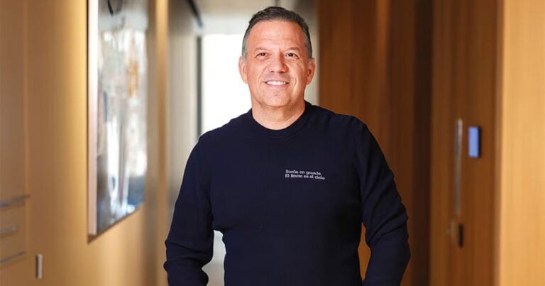 Las2orillas.co: Historias, voces y noticias de Colombia Smiling man in a navy sweater stands in a sunlit hallway with wood-paneled walls. - Las2orillas.co: Historias, voces y noticias de Colombia