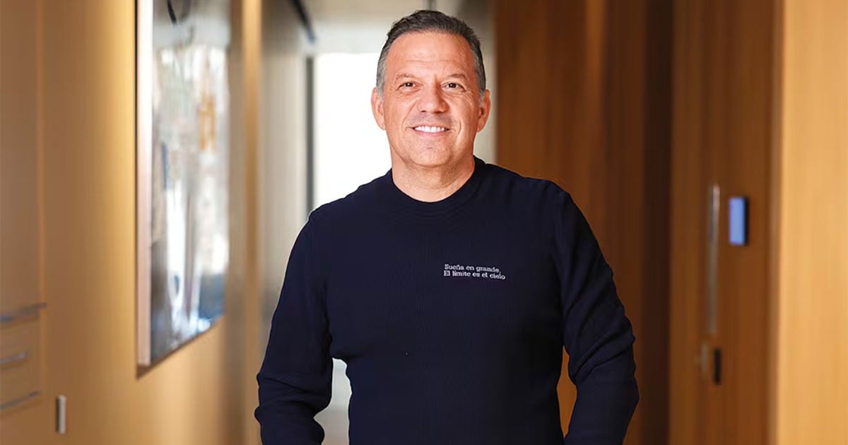 Smiling man in a navy sweater stands in a sunlit hallway with wood-paneled walls. - Un millonario pleito por fraude persigue en Nueva York al empresario y excandidato Santiago Botero