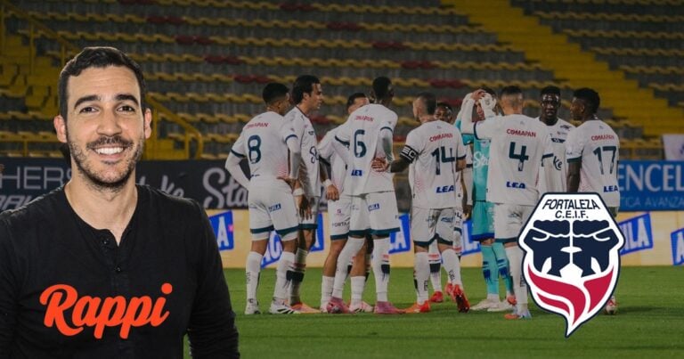 Las2orillas.co: Historias, voces y noticias de Colombia Smiling man in a black Rappi shirt stands in the foreground as a soccer team huddles on the field behind him, with a Fortaleza logo visible on the right. - Las2orillas.co: Historias, voces y noticias de Colombia