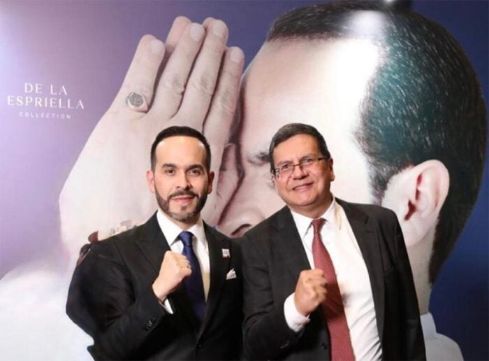 Así se escribió el Libro de la Verdad que De La Espriella entregó a la Fiscal con 126 escandalosos casos de la era Petro Two men in suits pose with fists raised at a fashion event, standing before a large poster of a model's face and the 'De La Esprilla Collection' text. - Así se escribió el Libro de la Verdad que De La Espriella entregó a la Fiscal con 126 escandalosos casos de la era Petro