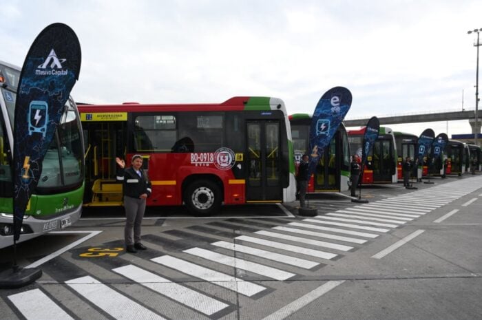 Los chinos en asocio con fabricantes de carrocerías caleños y brasileños hicieron 50 buses duales de Transmilenio Line of colorful city buses parked in a row at a bus depot, with a staff member waving at the camera and promotional banners nearby. - Los chinos en asocio con fabricantes de carrocerías caleños y brasileños hicieron 50 buses duales de Transmilenio