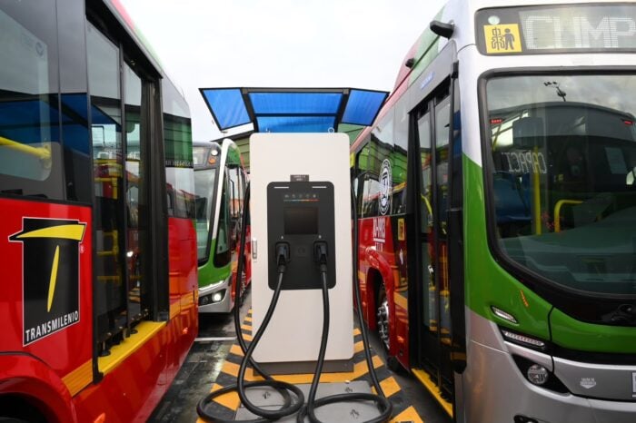 Los chinos en asocio con fabricantes de carrocerías caleños y brasileños hicieron 50 buses duales de Transmilenio Bus charging station in a bus depot surrounded by colorful city buses, with cables plugged into a tall white charging unit. - Los chinos en asocio con fabricantes de carrocerías caleños y brasileños hicieron 50 buses duales de Transmilenio